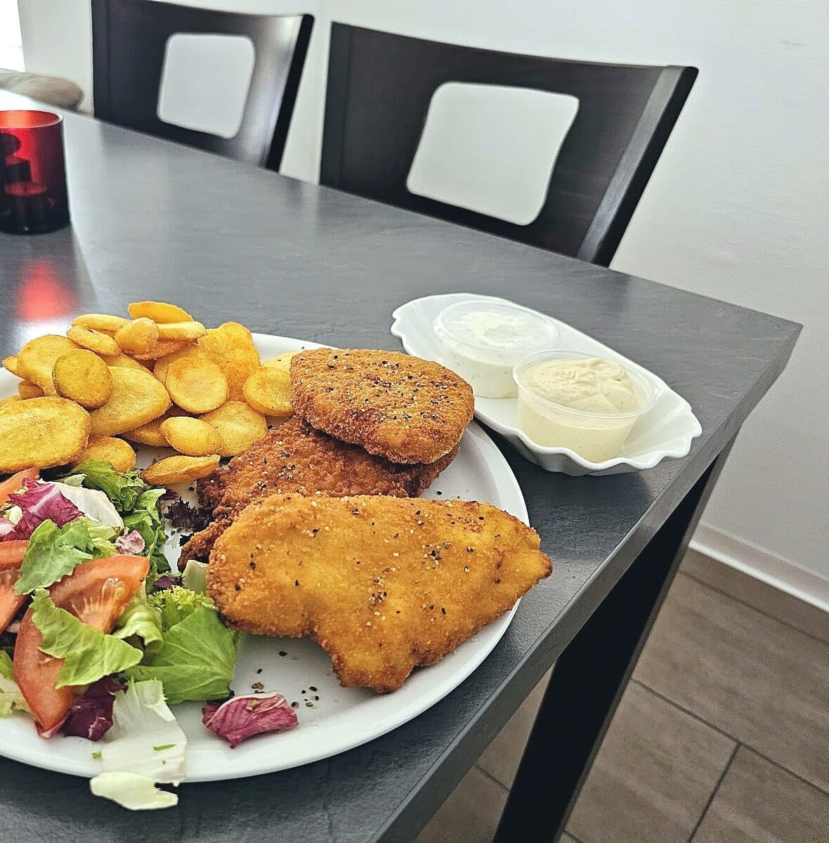 Drei panierte Schnitzel mit Kartoffelscheiben, Salat und zwei Dips auf einem Teller