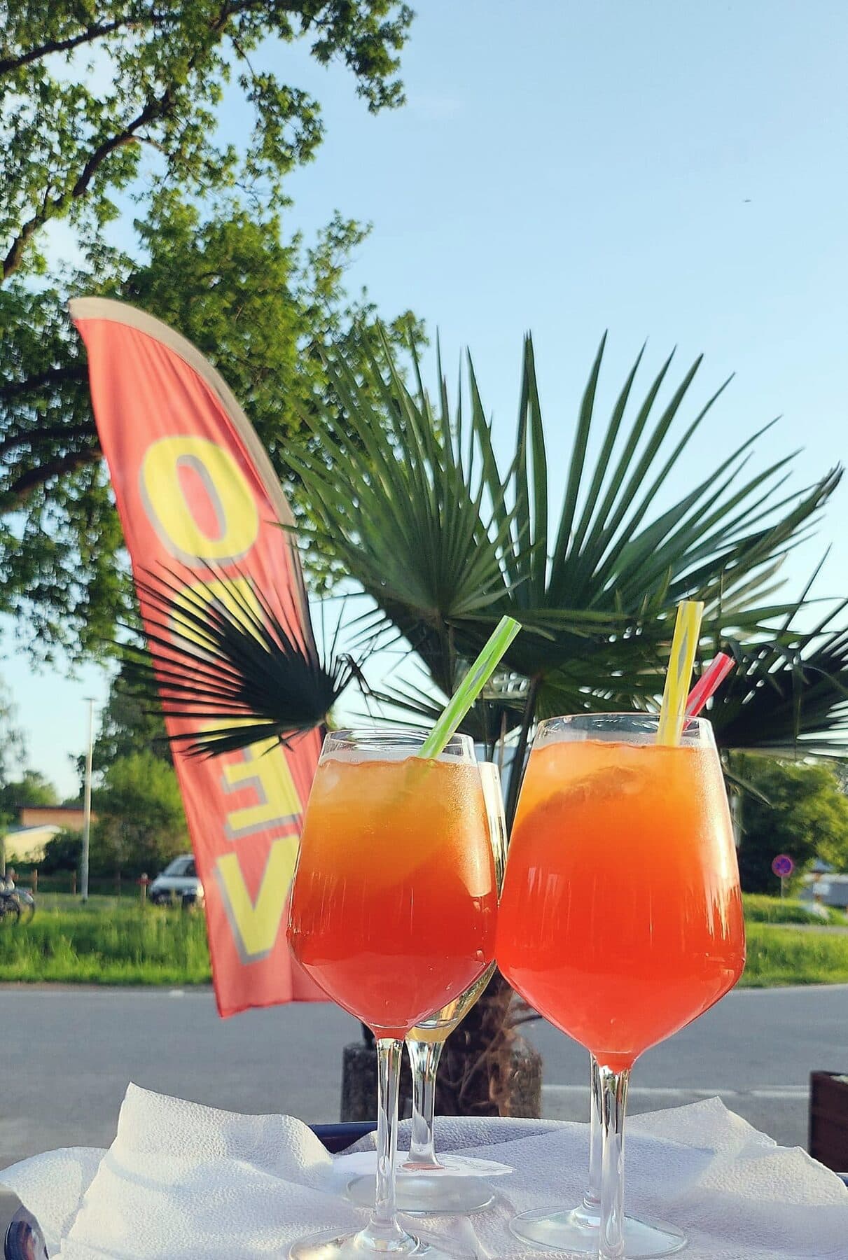 Orange Sommerdrinks in Weingläsern mit Strohhalmen auf der Terrasse, dahinter Palme und rote Fahne